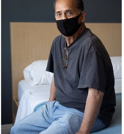 An older man wearing a mask sits on a bed, reflecting resilience and hope in his journey toward stable housing.