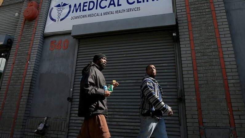 Two individuals walk past a medical clinic in downtown Los Angeles, highlighting access to health services for the community.