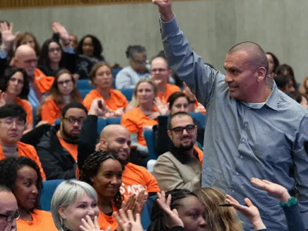 A community member passionately raises his fist during a meeting, surrounded by engaged attendees in orange shirts.