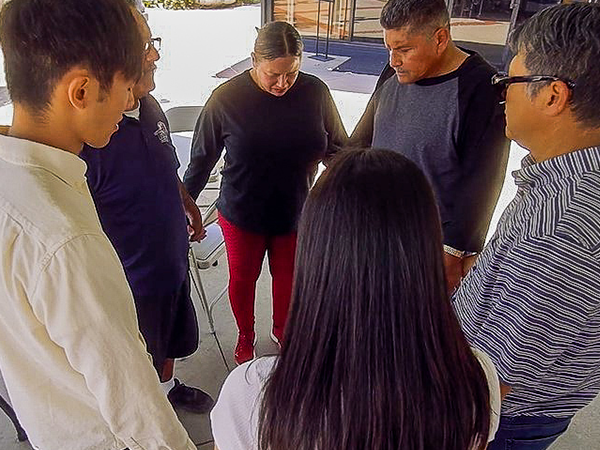 A group of six individuals stands in a circle, holding hands in a moment of prayer and support for homelessness solutions.