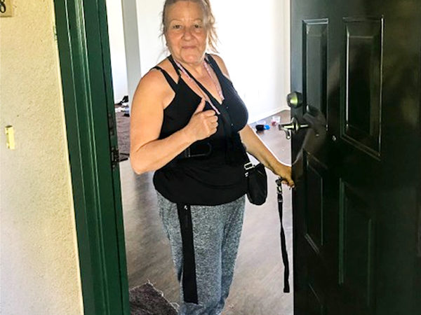 A woman smiles and gives a thumbs-up as she stands at the doorway of her new home, celebrating her transition to stable housing.