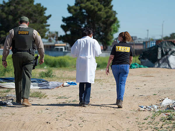 A sheriff and a healthcare worker walk alongside a community member, addressing health needs in a homeless encampment.