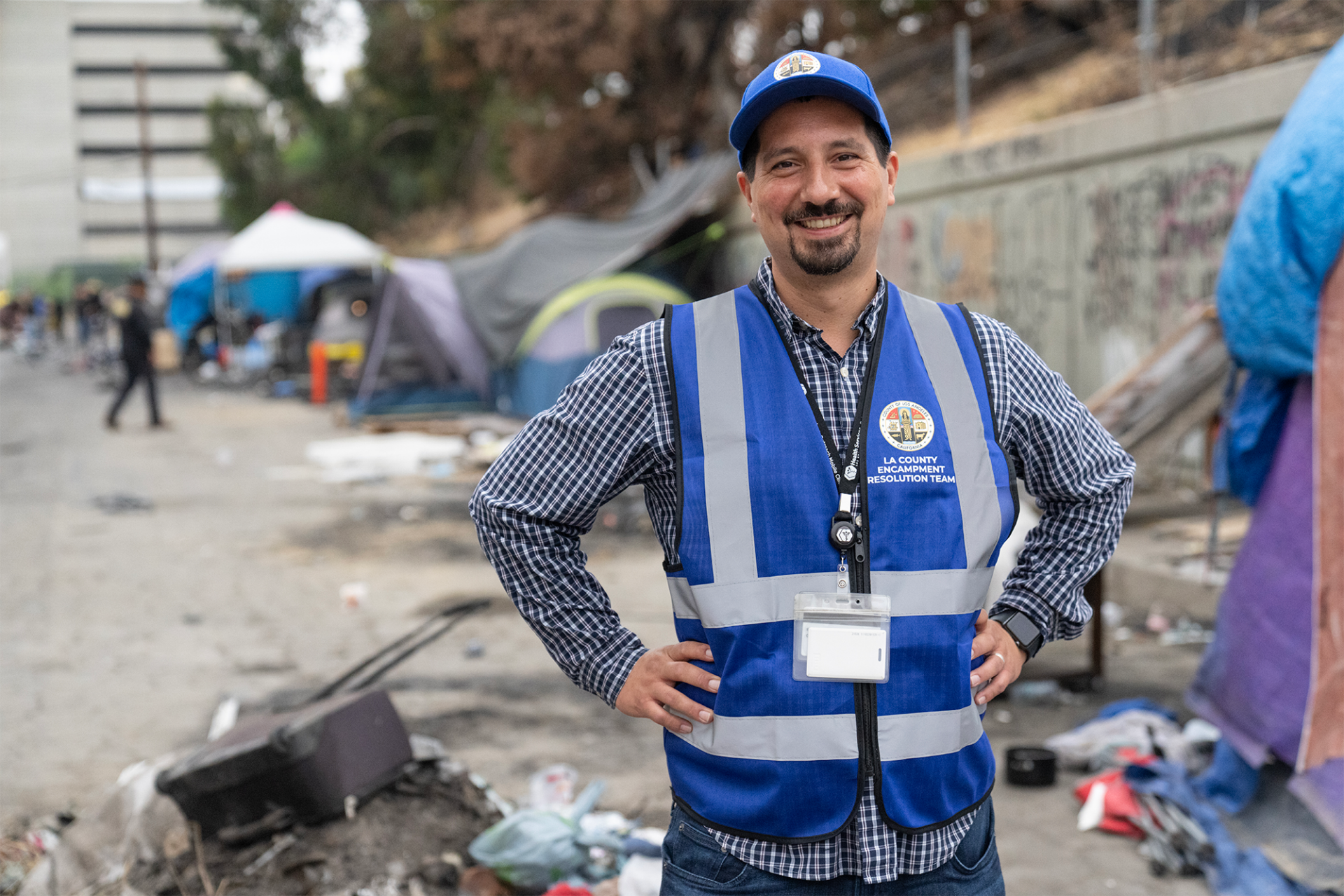 A smiling outreach worker in a vest stands confidently near a homeless encampment, embodying hope and support for the community.