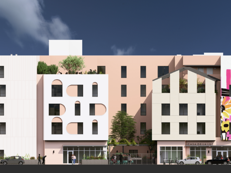 This image depicts a modern, colorful, multi-story building featuring a blend of architectural styles and materials. The façade includes various design elements: On the left, a minimalist white section with rectangular windows. A central portion with soft pink walls and arched windows. A section with beige brick-like patterns forming geometric cutouts. A striking mural on the right, showcasing vibrant floral artwork with sunflowers and a yellow sun against a pink-and-white background. The building is surrounded by greenery, including rooftop plants and ground-level landscaping. The entrance, labeled "Casa de la Luz," suggests it might serve as a residential or community-oriented structure.