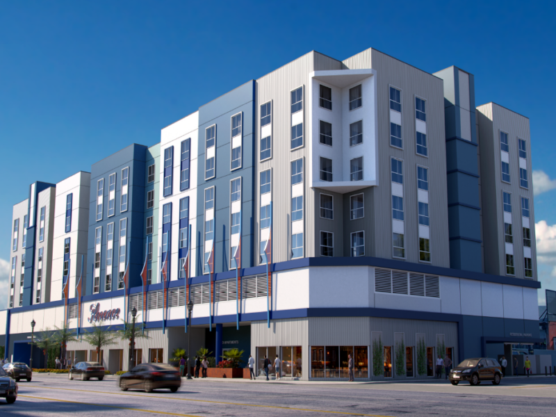 This image shows a contemporary mid-rise building with a sleek, urban design. The structure features: A façade dominated by vertical panels in shades of blue and white, complemented by evenly spaced windows. A central section with a recessed balcony area framed by clean, angular lines. A ground floor that appears to house commercial spaces, with large glass storefronts and signage for "Samaritan" prominently displayed. Multiple flags lining the perimeter of the building, adding a formal or institutional touch. Surrounding elements like parked cars, pedestrians, and a busy street, suggesting the building is situated in a bustling urban environment. The design reflects functionality and modern aesthetics, likely serving as a mixed-use or residential facility with community-oriented features.