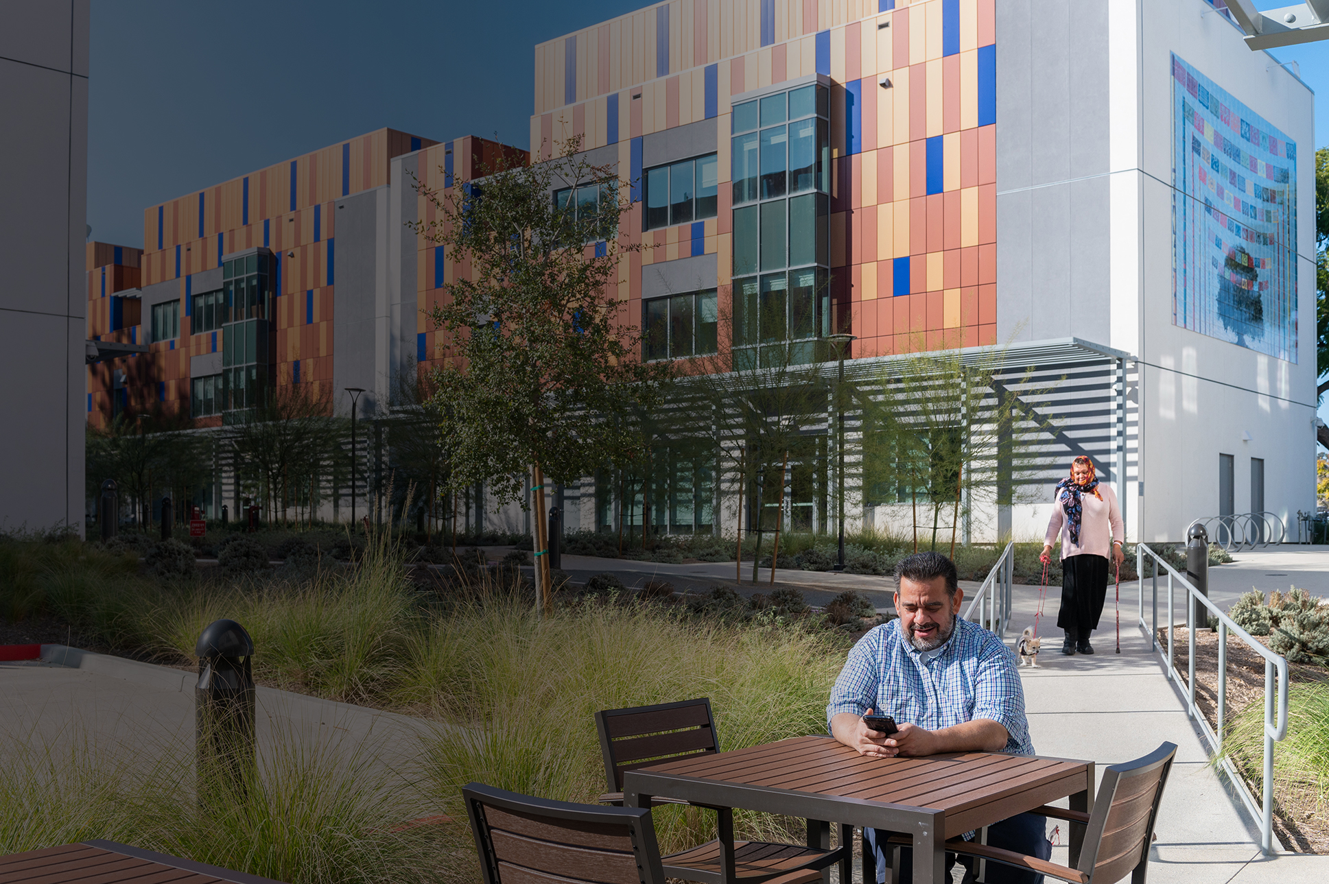 A man sits at a table using his phone, while a woman walks towards a colorful supportive housing building.
