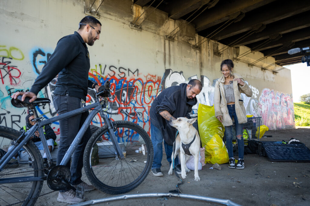 LAHSA outreach worker Marco Garcia, left, talks with Anthony and Karicha at their encampment before moving them to interim housing during a Pathway Home operation in South Gate, Dec. 10, 2025. (Photo by Michael Owen Baker)