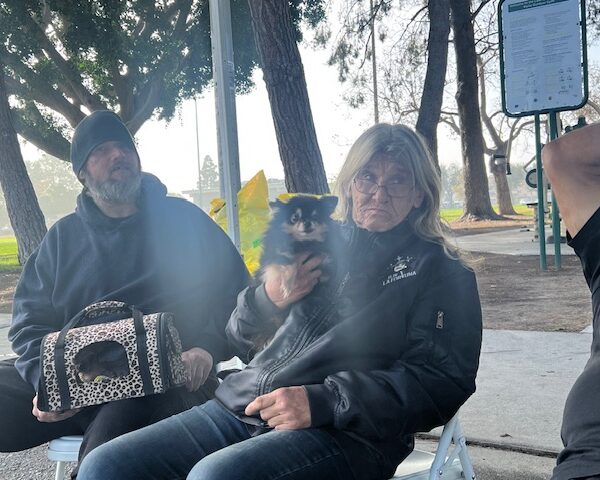 A man and woman with a dog in her arms sit on a bench outside.