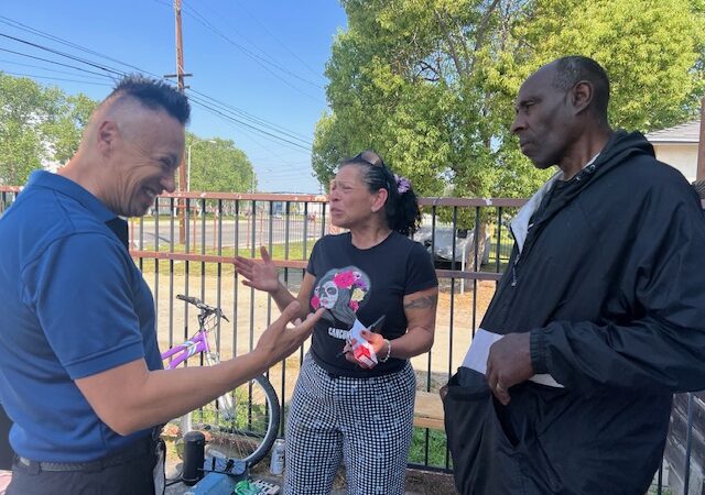 A man in a blue shirt smiles and engages with a woman in a black t-shirt and a man in a black sweatshirt.